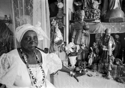 Uma foto em preto e branco de uma mulher negra a vovó maria Joanna do jongo da serrinha vestida de branco fumando uma cachimbo em frente a uma coleção de santos no seu terreiro