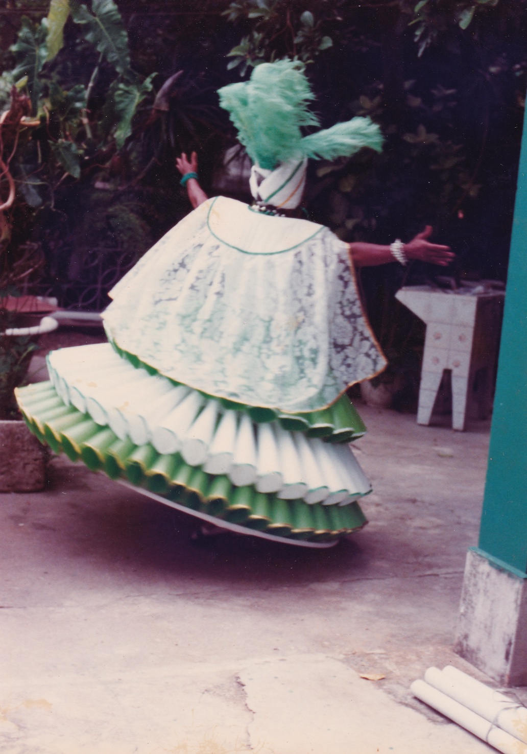 0000033 Em um quintal, Tia Maria, uma mulher negra, está de costas, com os braços abertos, com fantasia de baiana. Às costas do vestido, há uma pala branca, contornada em verde. Dela desce um tecido branco todo rendado, cobrindo ombros e costas. A saia rodada do vestido está inclinada, dando a sensação de que Tia Maria rodopiava com ele. Ela tem três camadas formada por tubos, semelhantes a cornetas. A primeira e a última são verdes, e a do meio é branca. Na cabeça, usa um turbante branco com cordões dourados e verdes. No topo, plumas verdes. Está com colares e pulseiras de bolas em prata, branco e verde.
