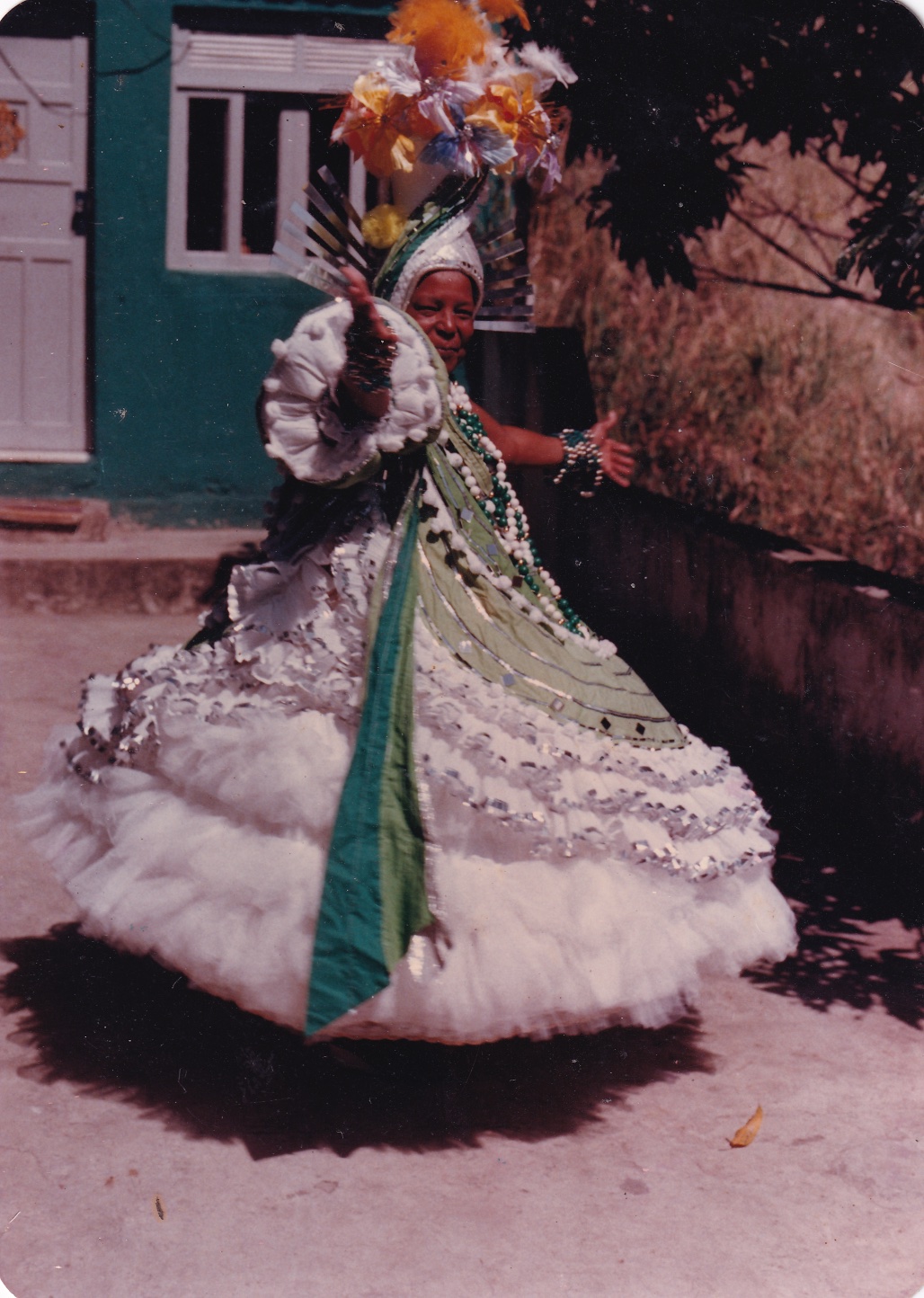 Tia Maria, uma mulher negra, está em um quintal, em frente a uma casa com porta e janela brancas. Ela está de perfil, com um sorriso nos lábios e com os braços abertos. Usa fantasia de baiana. O vestido, em verde e branco, desce em curvas, como uma espiral. Há faixas em verde claro, separadas por um cordão branco, e a saia é rodada em branco, em tecido fino. Todo ele é contornado por fitas prateadas. A manga, que vai até o cotovelo, é bufante. A parte interna, vista pelo braço estendido, é branca. Do ombro até embaixo, descem faixas em tons de verde com fitas pratas. Usa colares com grandes bolas em verde e branco, assim como as pulseiras. Na cabeça, há um adereço com arranjo de flores amarelas, brancas e azuis. Dele, descem faixas verdes, como as da lateral do vestido. Por trás, há um arco formado por tiras pratas.
