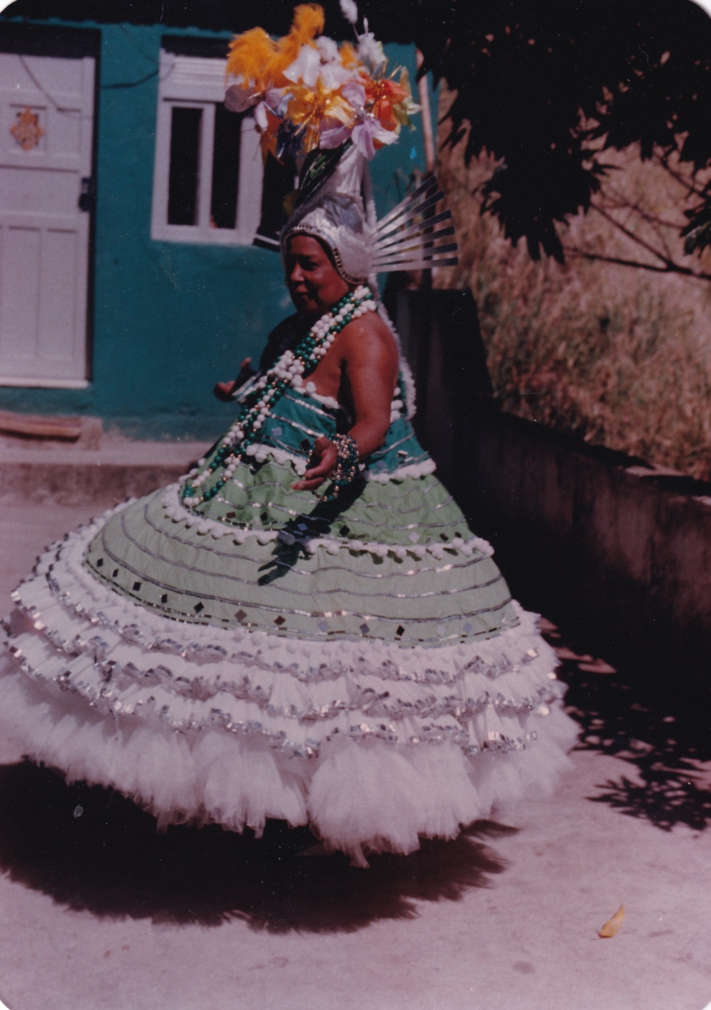 Tia Maria, uma mulher negra, está em um quintal, em frente a uma casa com porta e janela brancas. Ela está de perfil, com um sorriso nos lábios e com os braços abertos na altura da cintura. Usa fantasia de baiana. O vestido, em verde e branco, deixa o ombro à mostra e desce em curvas, como uma espiral. Inicia em verde escuro, depois há duas faixas em verde claro, separadas por um cordão branco, e por último, a saia rodada em branco, em tecido fino. Todo o vestido é contornado por fitas prateadas. O tronco dela é coberto por colares com grandes bolas em verde e branco, assim como as pulseiras que usa. Na cabeça, há um adereço com arranjo de flores amarelas, brancas e azuis. Por trás, há um arco formado por tiras pratas.