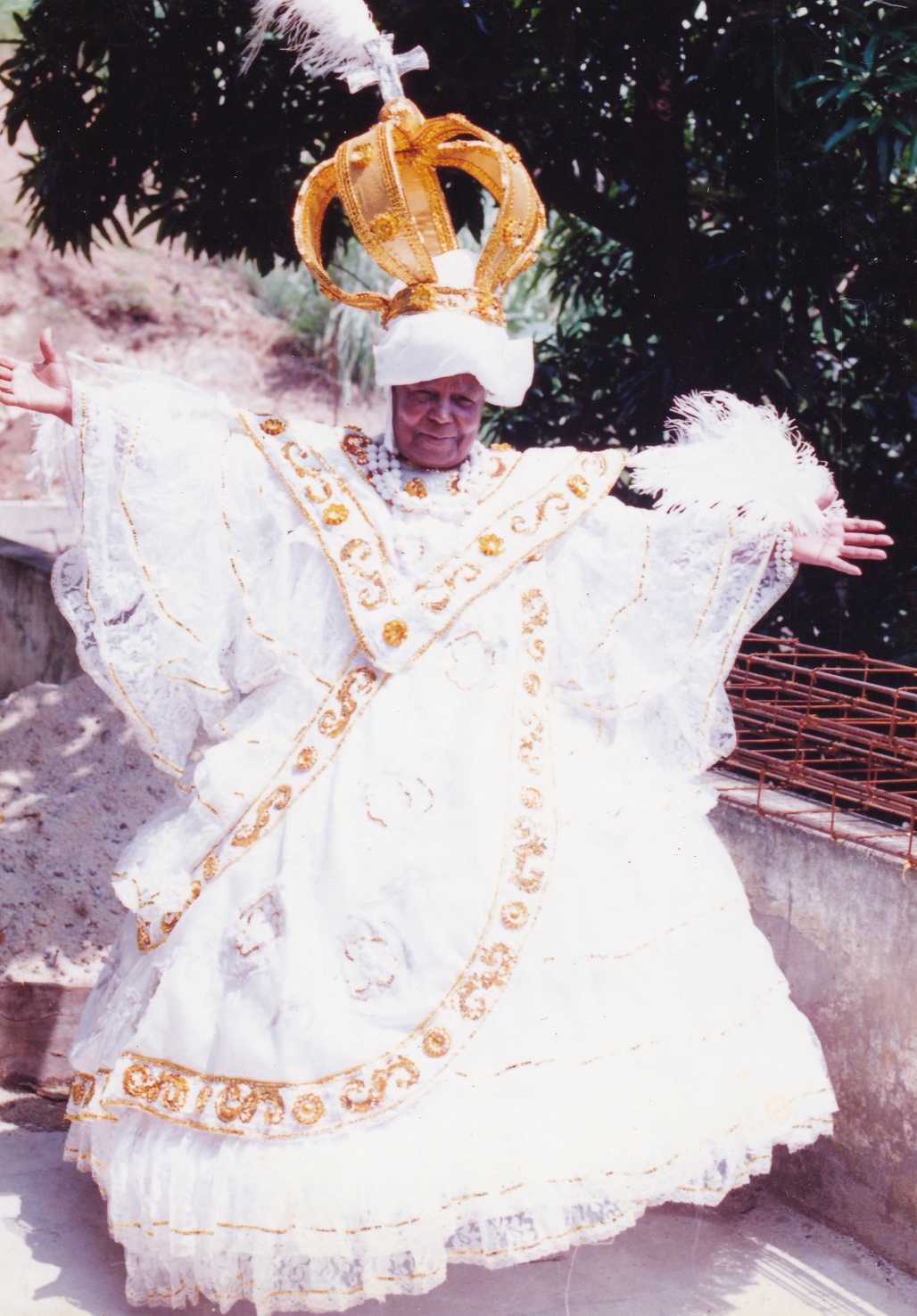 Em um quintal, à frente de uma árvore com folhas verdes, Tia Maria, uma mulher negra, está com fantasia de baiana. O vestido é branco, todo rendado, com símbolos dourados em ‘V’ à frente do peito e em linhas que passam pela saia rodada. Há também filetes dourados que contornam toda a fantasia. Ela está com os braços abertos, de forma que as mangas longas do vestido formam arcos, unindo-se à cintura. Acima dos ombros, há plumas brancas, na horizontal. Na cabeça, usa um turbante branco e acima dele uma enorme coroa dourada, com uma cruz prata e uma pluma branca no topo. Tia Maria usa colares e pulseiras de bolas brancas.