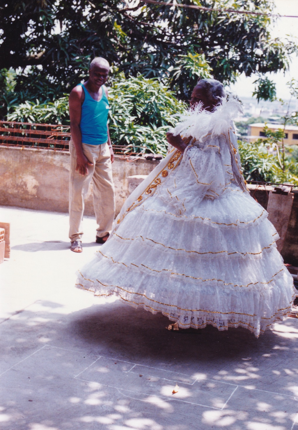 Tia Maria, uma mulher negra de cabelos grisalhos, está em um quintal, de perfil, fantasiada de baiana. O vestido é todo branco, rendado, com símbolos em dourado em uma tira à frente da roupa. Há também cordões dourados que contornam todas as camadas do vestido. Acima do ombro, há uma pluma branca, na horizontal. Perto de Tia Maria está um homem negro de cabelos grisalhos. Usa regata azul e calça bege. Olha para frente e sorri.
