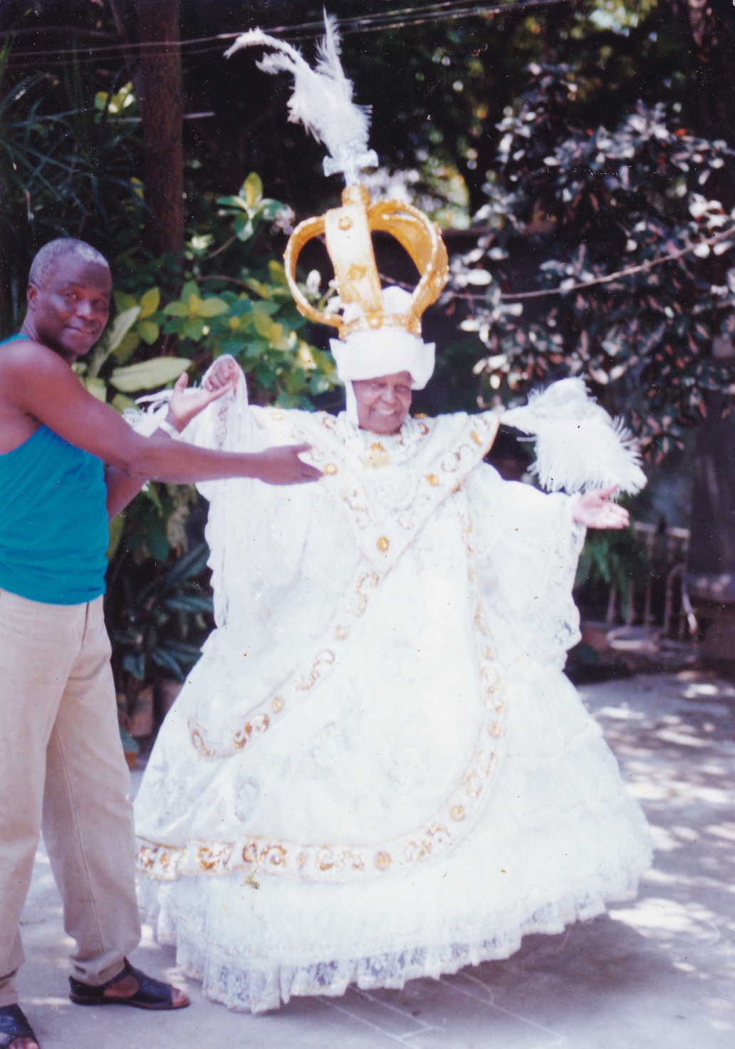 Tia Maria, uma mulher negra, está em um quintal com fantasia de baiana, ao lado de um homem negro de cabelos grisalhos. O vestido dela é branco, todo rendado, com símbolos dourados em ‘V’ à frente do peito e em linhas que passam pela saia rodada. Ela está com os braços abertos, de forma que as mangas longas do vestido formam arcos, unindo-se à cintura. Acima dos ombros, há plumas brancas, na horizontal. Na cabeça, usa um turbante branco e acima dele uma enorme coroa dourada, com uma cruz prata e uma pluma branca no topo. Está com colares e pulseiras de bolas brancas. O homem, que usa regata azul e calça bege, segura em uma das mãos de Tia Matia e aponta com o outro braço para ela. Os dois sorriem.