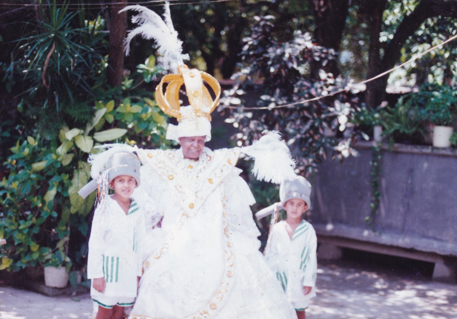 Tia Maria, uma mulher negra, está em um quintal, com fantasia de baiana, entre dois meninos também fantasiados. O vestido dela é branco, todo rendado, com símbolos dourados em ‘V’ à frente do peito e em linhas que passam pela saia rodada. Acima dos ombros, há plumas brancas, na horizontal. Na cabeça, usa um turbante branco e acima dele uma enorme coroa dourada, com uma cruz prata e uma pluma branca no topo. Está com colares de bolas brancas. Os meninos, um mais alto do que o outro, são pardos. As roupas, iguais, são compostas por camisas de mangas longas e bermudas brancas, com listas verdes em uma das golas, palas e bolsos, bem como nas barras das bermudas. Estão com uma espécie de capacete cinza, com plumas brancas no topo e fitilhos coloridos na lateral. Eles olham para frente. Tia Maria sorri.
