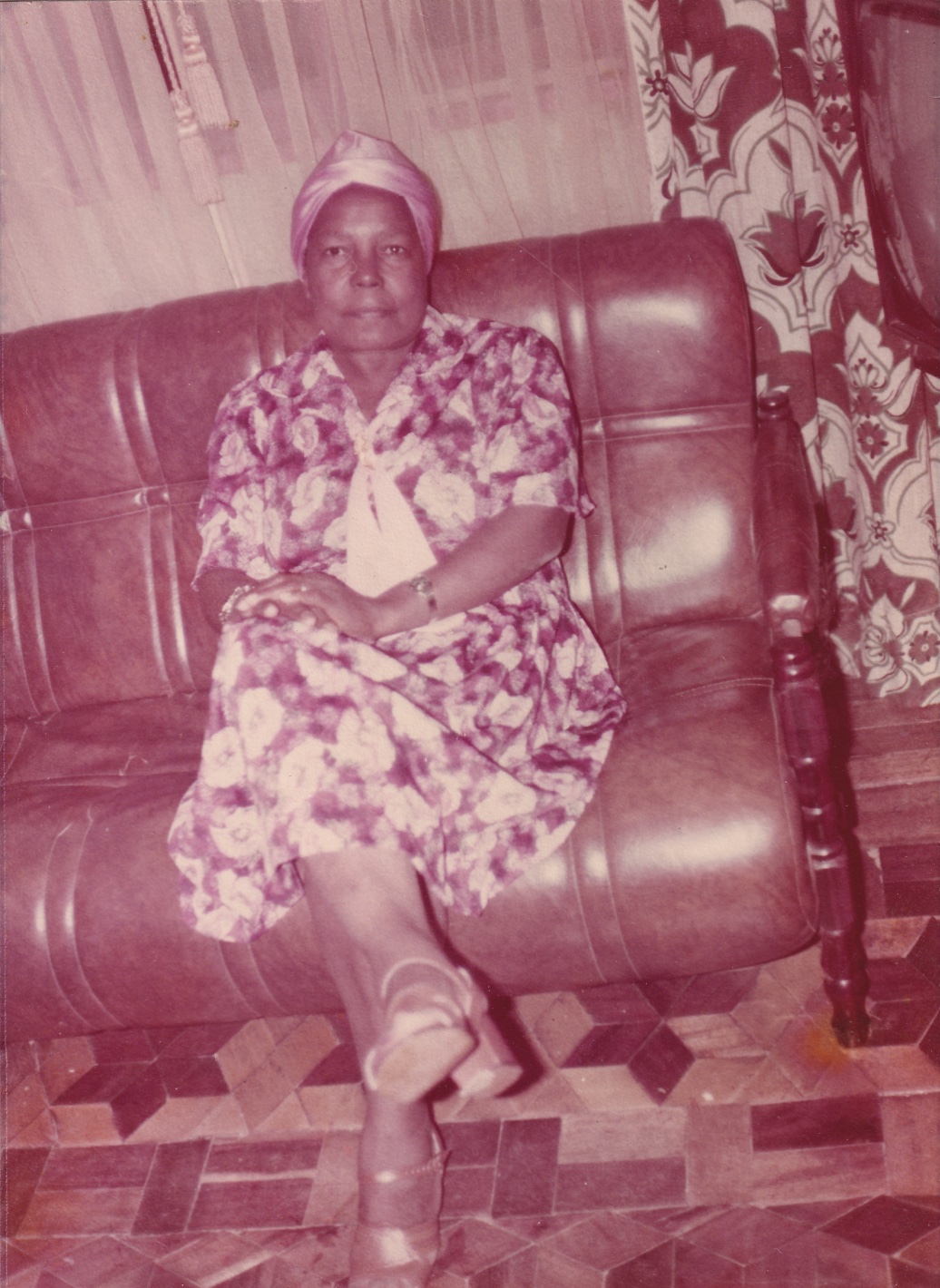 Em foto ligeiramente inclinada, Tia Maria está sentada em um sofá marrom, à frente de cortinas brancas fechadas. Ela é uma mulher negra. Olha para frente, com as pernas cruzadas e as mãos sobre a coxa. Usa turbante rosa claro e vestido florido longo, de mangas curtas, tendo à frente um lenço branco amarrado, na vertical. Em um dos pulsos usa pulseira dourada e no outro um relógio da mesma cor. Nos pés, sandálias de salto, também douradas. O chão da sala é em tacos de madeira, formando desenhos.