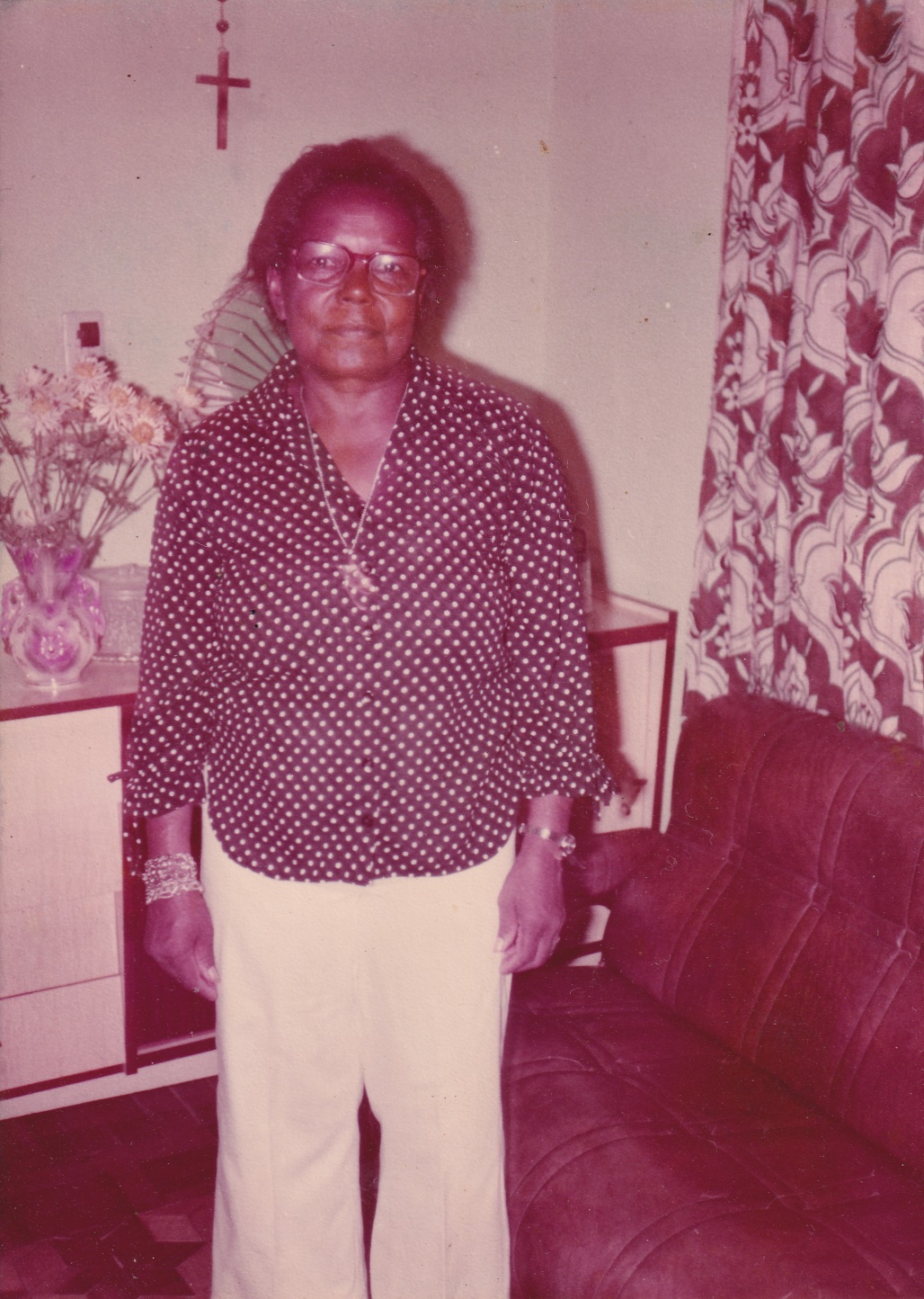 Tia Maria, uma mulher negra de cabelos pretos penteados para trás, está em pé em uma sala. Olha para frente. Usa óculos de grau quadrados e está vestida com camisa de mangas longas preta com poás brancos, e calça branca. Usa cordão com pingente prata. As mangas da camisa estão dobradas, deixando à mostra pulseiras douradas e um relógio da mesma cor. Atrás dela há um crucifixo de madeira pendurado na parede e um armário de madeira bege, à meia altura, com um vaso com flores. Ao lado há cortinas estampadas e um sofá marrom.