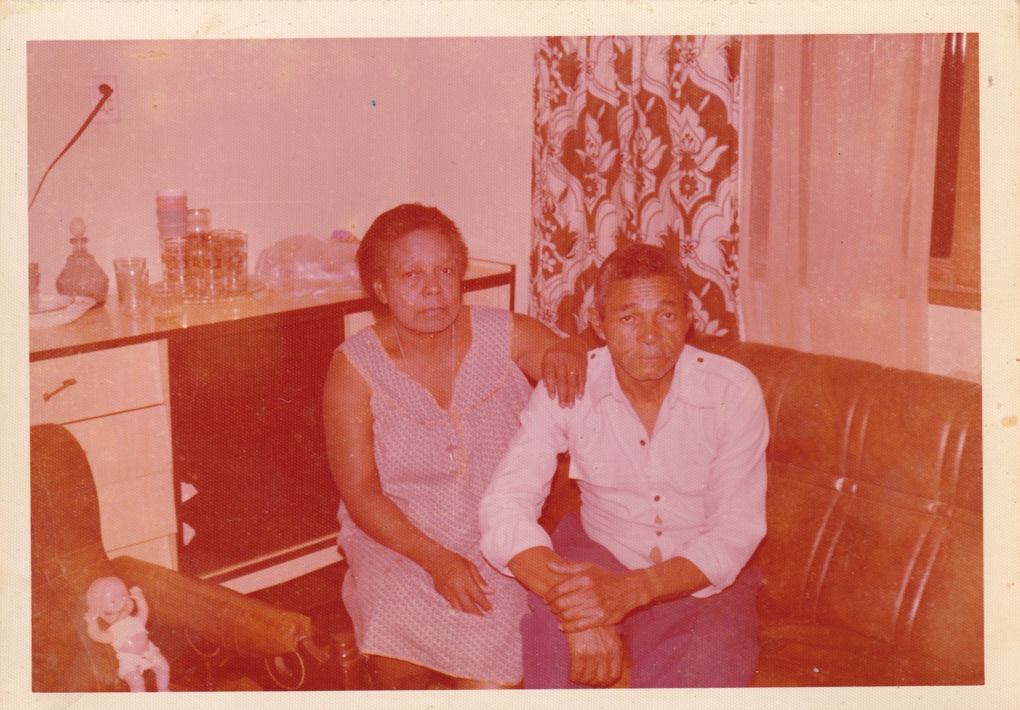 0000047 Sentados em um sofá marrom em uma sala, estão Tia Maria e seu marido Ivan. Ela é uma mulher negra de cabelos grisalhos curtos e cheios. Usa vestido com alças, quadriculado em azul claro e branco. Está com uma das mãos apoiada no ombro de Ivan. Ele é um homem negro de cabelos grisalhos. Usa camisa de mangas longas branca, com os punhos dobrados, e calça azul. Ambos olham para frente. Na parede ao lado há um armário de madeira bege, à meia altura, com um jogo de copos estampados em dourado acima dele. À frente, há uma poltrona, como o sofá, com uma boneca de plástico em cima. Atrás deles, cortinas estampadas e brancas.