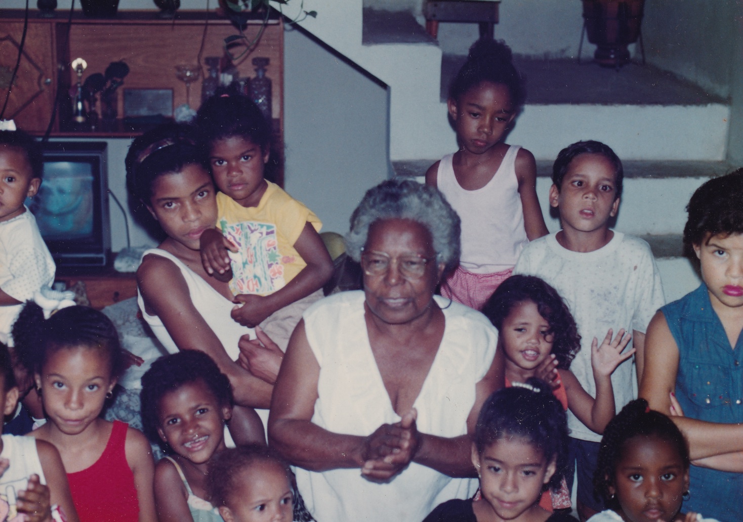 Tia Maria, uma senhora de pele negra e de cabelos grisalhos curtos e cheios, está em uma sala rodeada por crianças, meninos e meninas, todos de peles negras. Elas olham para frente e algumas estão com as mãos posicionadas como se batessem palmas, inclusive Tia Maria. Ela usa óculos de grau quadrados e blusa branca, sem mangas. As crianças estão com roupas coloridas. Atrás deles há uma estante com uma tv e os degraus de uma escada que sobe.