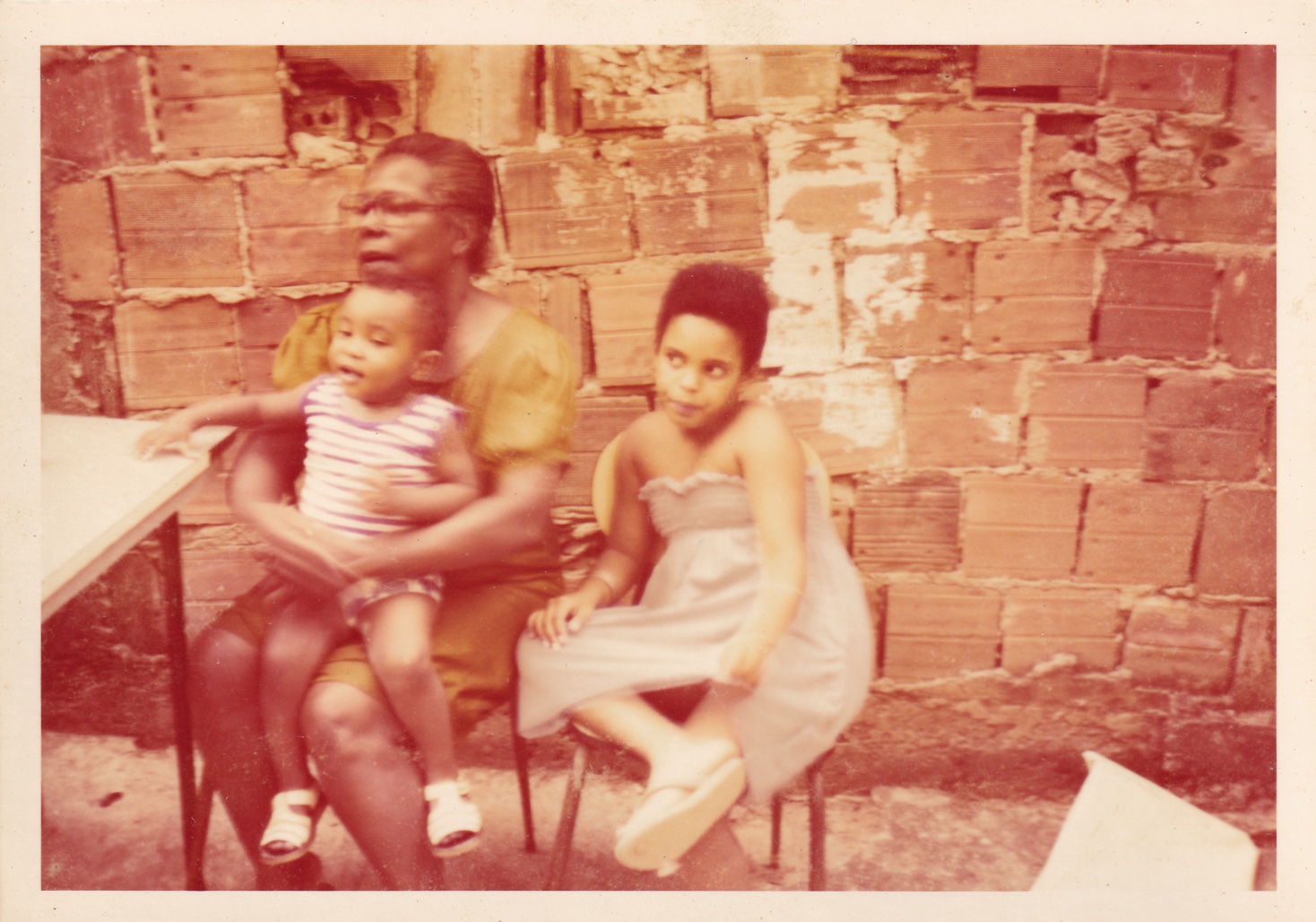 À frente de uma parede com tijolos aparentes, estão sentados em cadeiras, perto de uma mesa, Tia Maria, com um menino no colo, e uma menina. Tia Maria é negra de cabelos grisalhos penteados para trás. Usa óculos de grau quadrados e vestido verde. O menino, sentado no colo dela, é negro de cabelos castanhos. Usa regata listrada e short. A menina, na cadeira ao lado, é negra de cabelos pretos curtos. Usa vestido lilás. Todos estão de frente, olhando para o lado. Tia Maria está com a boca parcialmente aberta, como se falasse.