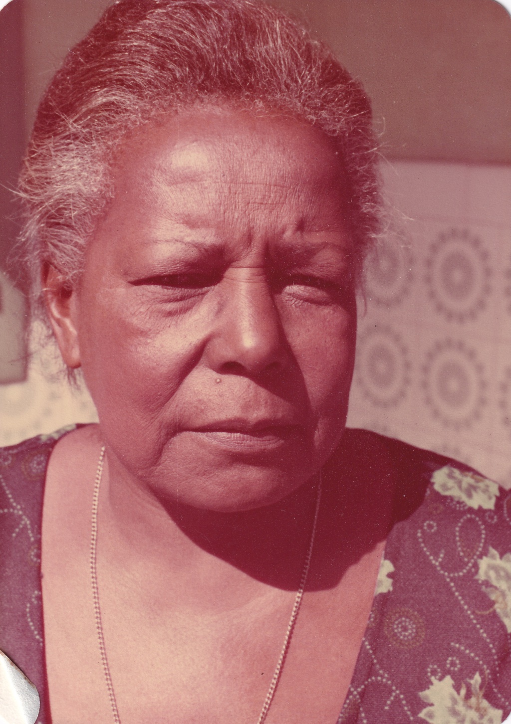 Tia Maria, uma senhora negra de cabelos grisalhos penteados para trás, é vista do busto para cima à frente de uma parede com azulejos brancos e cinzas, à meia altura. O rosto dela está no sol, de forma que olha para frente com as sobrancelhas franzidas. Usa blusa azul com estampas de folhas verdes, decotada em ‘V’, e cordão prata ao pescoço.