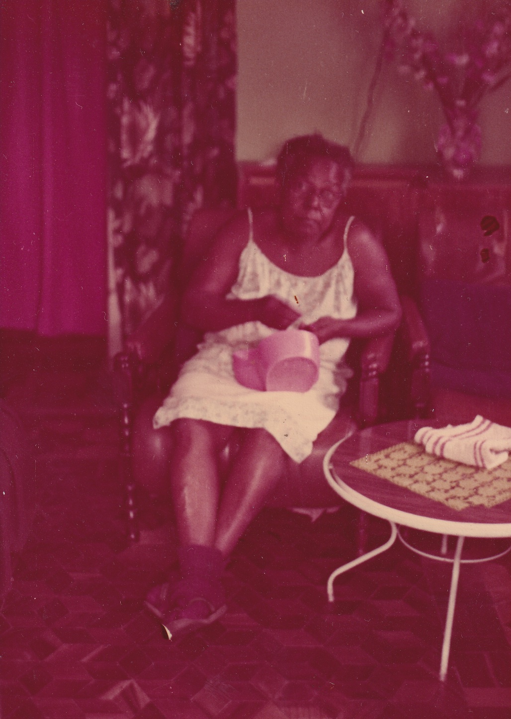 Na foto, um pouco desfocada, Tia Maria está sentada em uma poltrona marrom em uma sala. Ela é uma senhora de pele negra e de cabelos grisalhos curtos e cheios. Usa vestido de alças branco. Ela olha para frente e está com as mãos acima de um escorredor de arroz rosa, que está no colo dela. Os pés, calçados com botinhas, estão cruzados. Perto dela há uma mesa de centro redonda, com toalhas em cima. Ao fundo, cortinas estampadas e um armário à meia altura com um vaso com flores. O chão é em tacos de madeira, que formam desenhos.