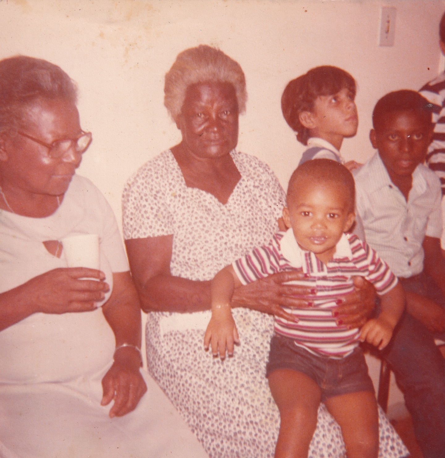 Em foto amarelada pelo tempo, estão, sentadas lado a lado à frente de uma parede branca, Tia Maria, uma senhora e crianças. À esquerda está Tia Maria, uma senhora negra de cabelos grisalhos penteados para trás. Ela usa óculos de grau quadrados, vestido claro e segura um copo plástico. Olha para uma criança que está sentada no colo da irmã, ao lado dela. Ela é uma senhora negra de cabelos brancos curtos e cheios. Usa vestido branco com pequenas estampas floridas. O menino que segura é negro, de cabelos escuros rentes à cabeça. Usa camisa polo listada e bermuda. Olha para frente, com a boca aberta, esboçando um sorriso. Ao lado, estão um menino branco, um menino negro, e outra pessoa vista parcialmente.