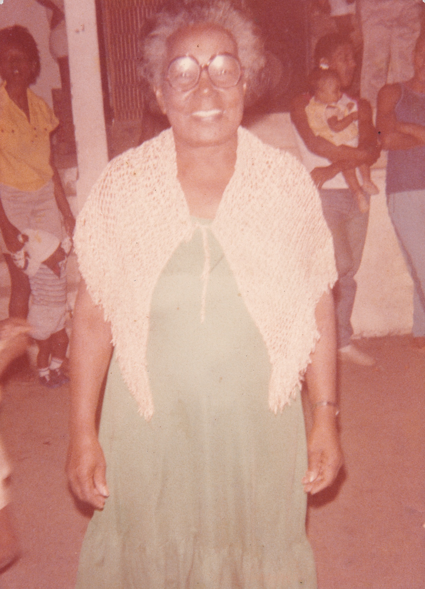 0000056 Em foto desbotada pelo tempo, Tia Maria está em uma espécie de salão, próxima a outras pessoas. Ela é uma senhora de pele negra, de cabelos grisalhos curtos e cheios. Está com óculos de grau redondos. Olha para frente com um largo sorriso no rosto. Usa vestido verde, com um chale tricotado em cor clara sobre os ombros e à frente do busto.