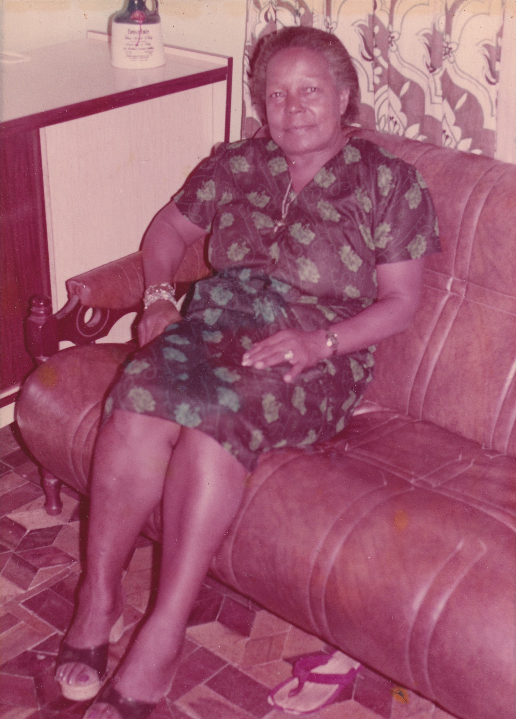 Tia Maria, uma mulher negra de cabelos grisalhos penteados para trás, está sentada em um sofá marrom em uma sala. Ela olha para frente, com um sorriso nos lábios. Usa vestido preto com estampas de flores verdes, de mangas curtas, um pouco abaixo dos joelhos. Está com um cordão ao pescoço e com as mãos apoiadas nas coxas. Usa, em um dos pulsos, pulseiras pratas e douradas. No outro, um relógio dourado e anel prata em um dedo. Está com tamancos pretos. Ao lado dela há um armário em madeira bege e marrom, à meia altura, e atrás do sofá, cortinas estampadas.