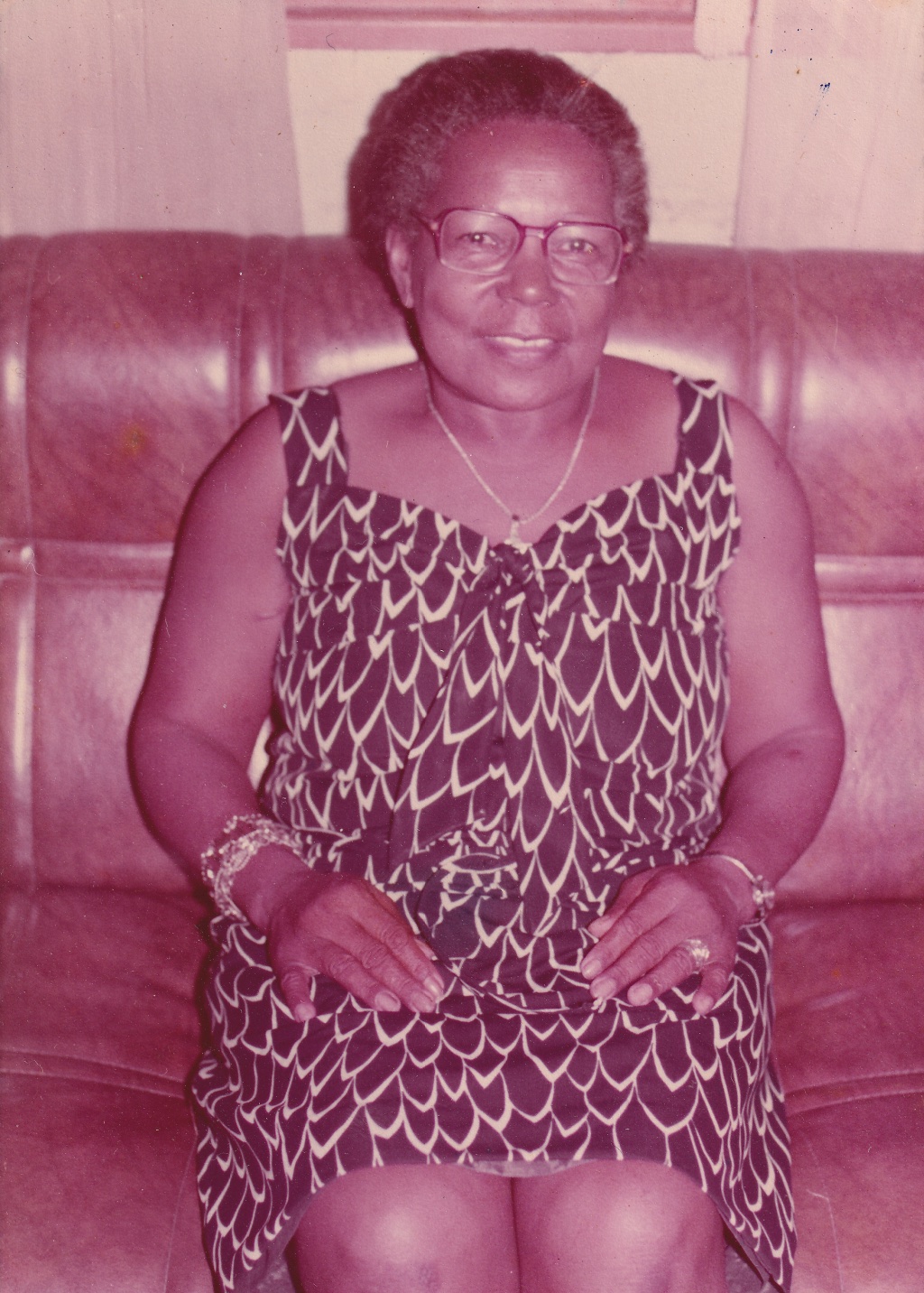 Tia Maria, uma mulher negra de cabelos grisalhos penteados para trás, está sentada em um sofá marrom em uma sala. Ela é vista dos joelhos para cima. Usa óculos de grau quadrados, olha para frente e sorri. Está com vestido de alças preto, com traços brancos, imitando folhas. Usa, cordão prata ao pescoço. Em um dos pulsos, pulseiras pratas e douradas. No outro, um relógio dourado e anel prata em um dedo.