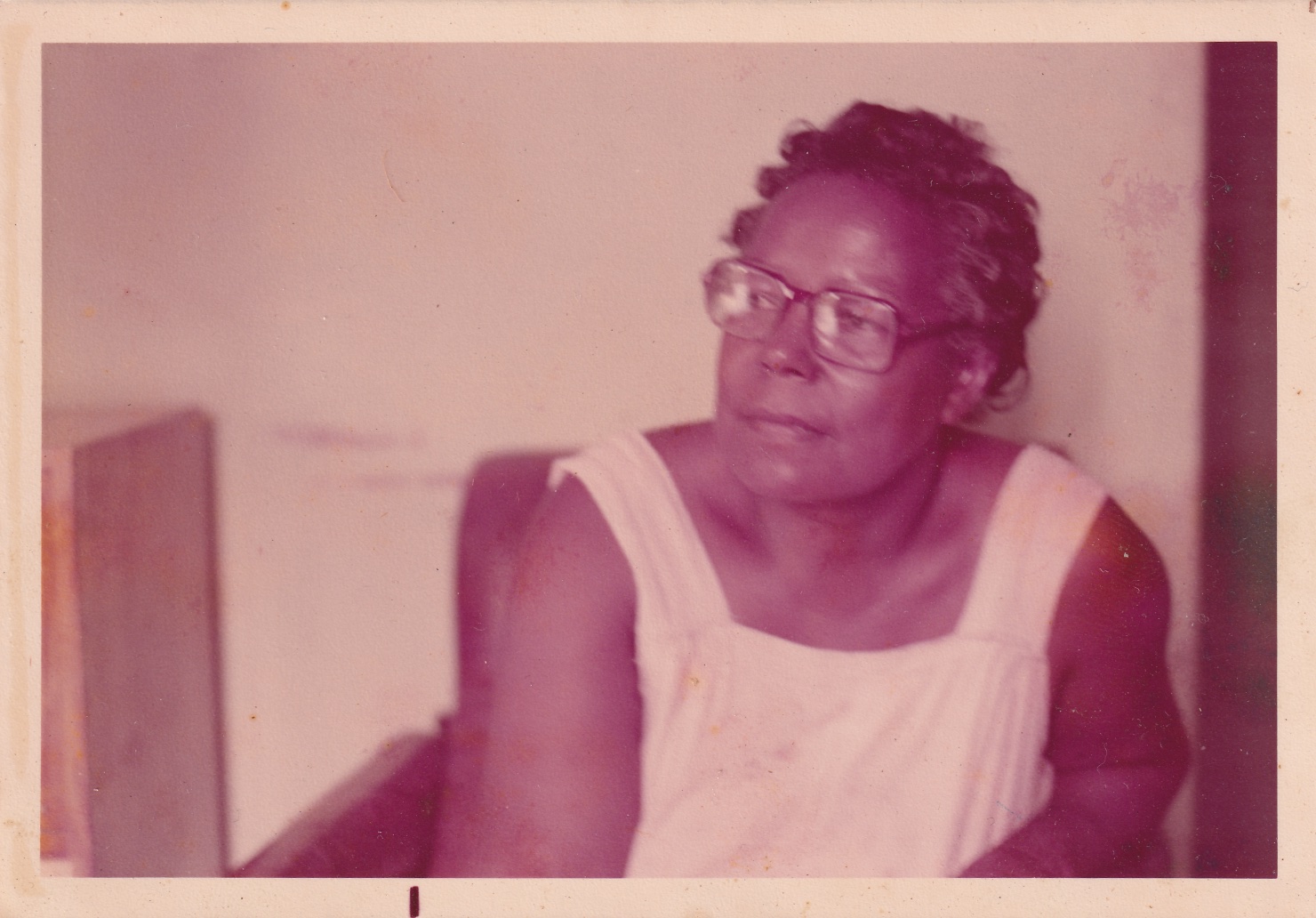 0000059 Em foto amarelada, Tia Maria está sentada em uma poltrona, à frente de uma parede branca. Ela é uma mulher negra de cabelos grisalhos cacheados curtos e usa óculos de grau quadrados. É vista da cintura para cima. Está de frente, com o rosto de perfil, olhando para o lado. Usa roupa branca, de alças.