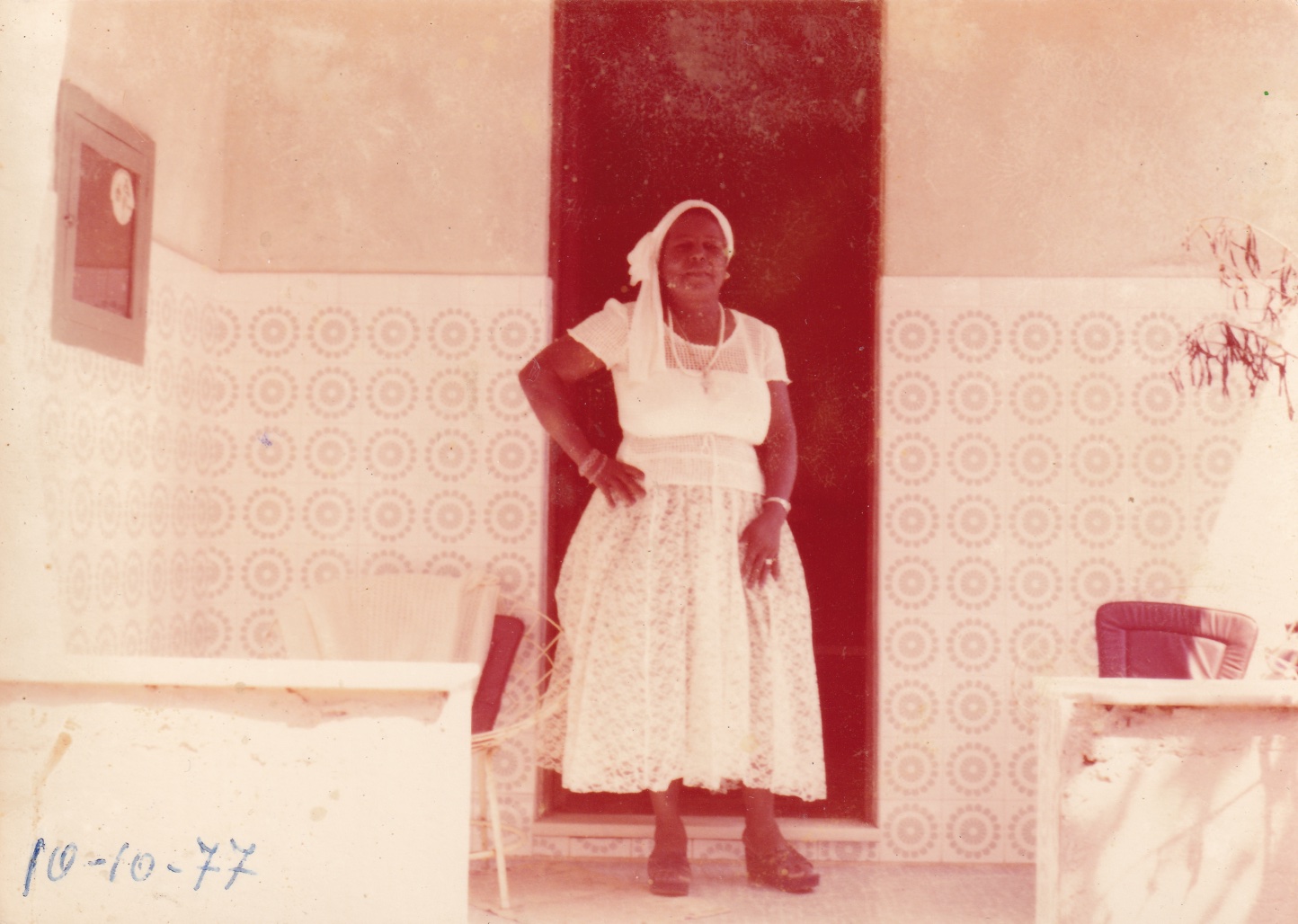 Em foto desbotada, Tia Maria está em pé em uma varanda, à frente de uma porta marrom fechada. Ela é uma mulher negra e usa roupas brancas: vestido rendado com mangas curtas e lenço à cabeça, que cai acima de um ombro. Ela olha para frente e está com uma das mãos na cintura. Usa colar, pulseiras, relógio, anel, e sandálias com salto plataforma. A varanda tem azulejos à meia altura, brancos com estampas cinzas. Os patamares são brancos. Nela, há cadeiras com estofados brancos e vermelhos. No rodapé da foto, está escrito à caneta: 10-10-77.