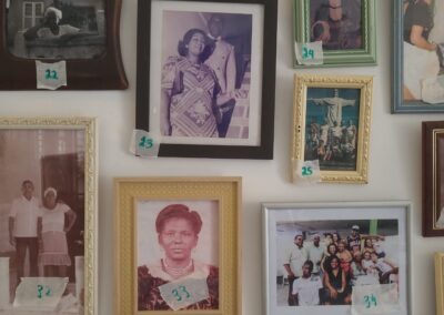 Em uma parede, há fotos de família emolduradas de vários tamanhos, cada uma identificada com pedaços de fita adesiva numerados. As imagens mostram pessoas sozinhas e em grupos, tanto em poses arranjadas quanto em momentos espontâneos, em diferentes ambientes e épocas.
