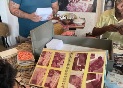 Uma pessoa segura um álbum de fotos aberto sobre uma mesa com toalha de renda. Outras duas pessoas estão organizando papéis; fotos antigas e retratos de família cobrem a parede ao fundo.