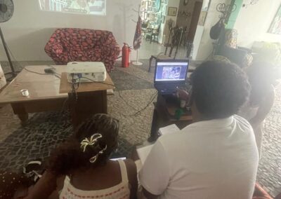 Duas pessoas estão sentadas em um sofá, usando um laptop que projeta uma imagem na parede de uma sala aconchegante e bem iluminada, com piso de pedra, móveis de madeira e decoração colorida.