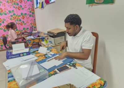 Duas pessoas trabalham em mesas cobertas por toalhas de mesa com estampa floral. A pessoa em primeiro plano corta papel com uma régua e um tapete de corte, enquanto a outra digita em um laptop ao fundo. As paredes têm decorações florais coloridas.