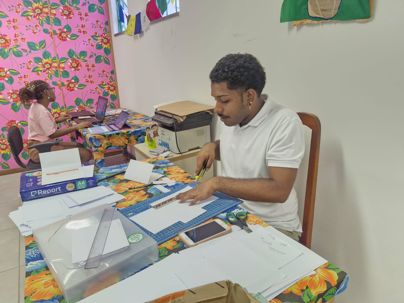 Duas pessoas trabalham em mesas cobertas por toalhas de mesa com estampa floral. A pessoa em primeiro plano corta papel com uma régua e um tapete de corte, enquanto a outra digita em um laptop ao fundo. As paredes têm decorações florais coloridas.