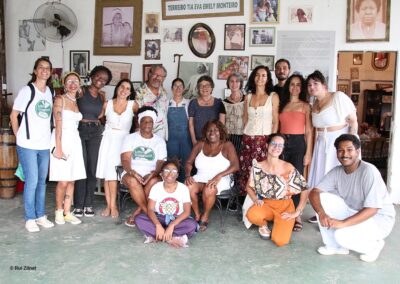 Um grupo de pessoas, sorrindo e posando juntas em um espaço interno decorado com fotos emolduradas e obras de arte na parede atrás delas. Algumas estão em pé, enquanto outras estão sentadas ou ajoelhadas na frente.