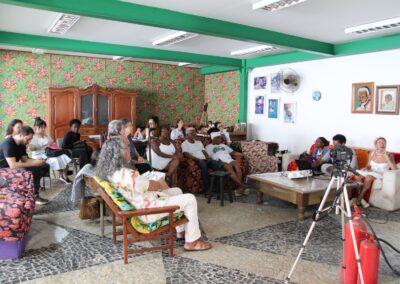 Um grupo de pessoas está sentado em sofás e cadeiras em uma sala colorida com papel de parede floral, observando ou ouvindo atentamente. Uma câmera montada em um tripé está posicionada à frente delas. Obras de arte estão penduradas na parede.