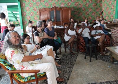 Um grupo diversificado de pessoas está sentado em cadeiras e sofás em uma sala decorada com cores vivas, observando atentamente algo que está fora do quadro.