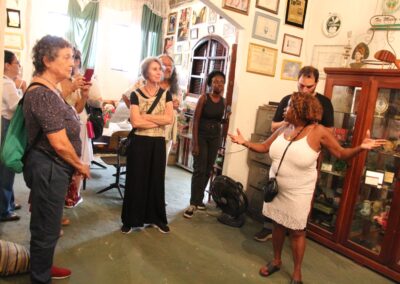 Uma mulher vestida com um vestido branco gesticula enquanto fala para um grupo de pessoas dentro de uma sala repleta de fotos emolduradas, prêmios e móveis antigos. O grupo ouve atentamente.