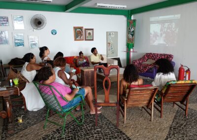 Um grupo de pessoas está sentado em uma sala aconchegante, assistindo a um vídeo projetado na parede. O espaço tem cadeiras de madeira, obras de arte nas paredes, um piso estampado e um ambiente descontraído.
