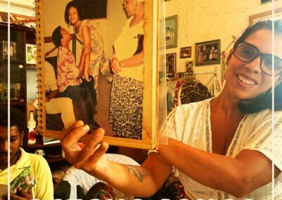 Uma mulher sorri enquanto segura uma foto emoldurada de dois idosos dançando. O fundo mostra uma sala colorida com enfeites. O texto diz: “Acervo Jongo da Serrinha” e “Centro de Memória Jongo da Serrinha”.