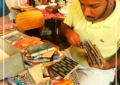 Um homem está sentado à mesa, examinando fotos e cartões-postais antigos. Vários objetos coloridos e itens de recordação estão espalhados ao redor. A legenda diz: “Acervo Jongo da Serrinha” e há um logotipo no canto superior direito.