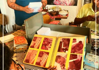 Três pessoas examinam fotografias e documentos antigos em uma mesa, com um grande álbum de fotos aberto em primeiro plano. A legenda da imagem diz: “ACERVO JONGO DA SERRINHA — Centro de Memória Jongo da Serrinha”.