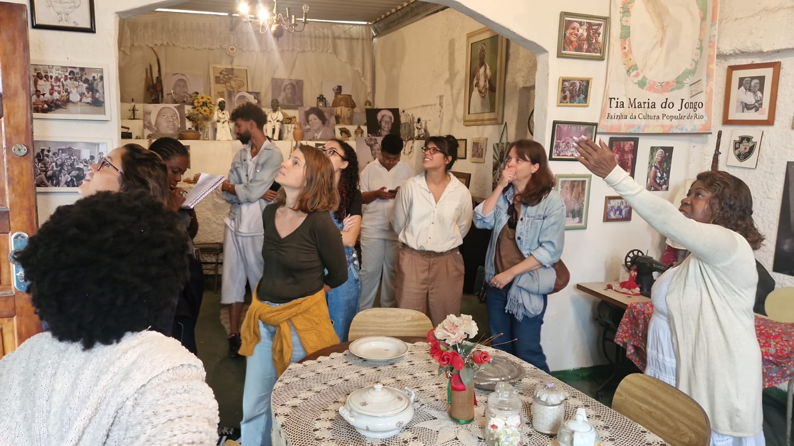 Um grupo de pessoas está de pé, olhando ao redor de uma sala aconchegante, decorada com fotos, obras de arte e mesas cobertas com rendas, enquanto uma mulher à direita gesticula e fala, possivelmente conduzindo uma visita guiada ou compartilhando informações.