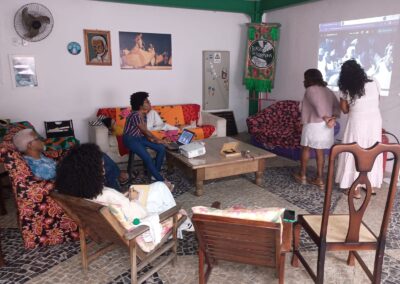 As pessoas estavam reunidas em uma sala com uma tela de projeção exibindo um vídeo, sentadas em cadeiras e sofás em frente a uma mesinha de centro baixa.