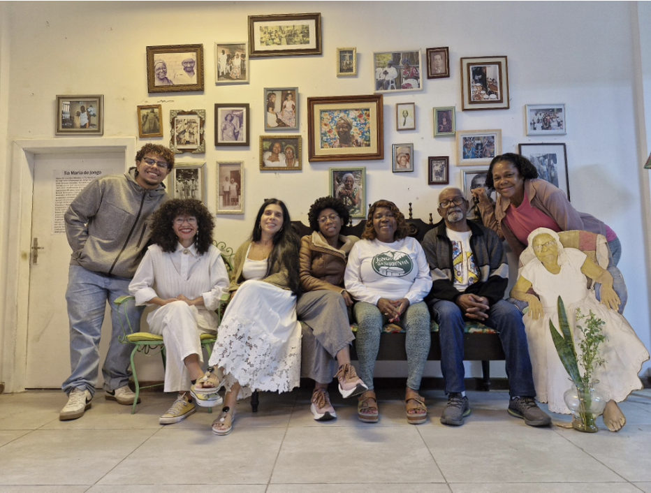 Um grupo de sete pessoas sentadas em um banco em frente a uma parede coberta de retratos emoldurados.
