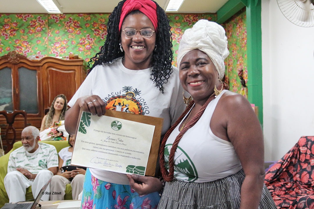Duas mulheres sorrindo, enquanto uma delas segura um certificado emoldurado; ao fundo, um homem e uma mulher est&atilde;o sentados em uma sala com papel de parede estampado com motivos florais.