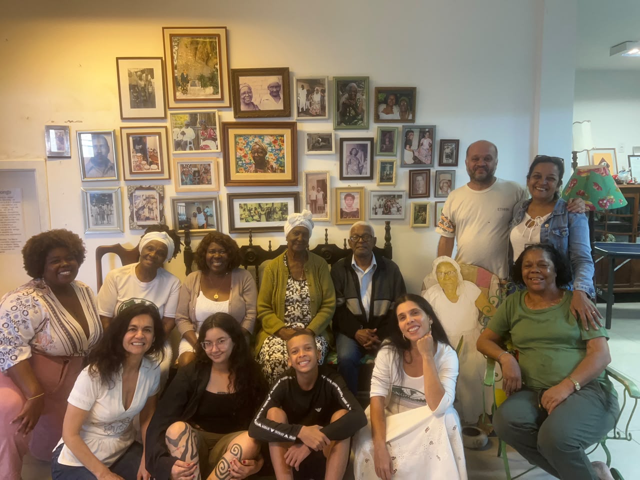 Um grupo diversificado de onze pessoas se reuniu para uma foto em uma sala com uma grande cole&ccedil;&atilde;o de fotografias emolduradas na parede.