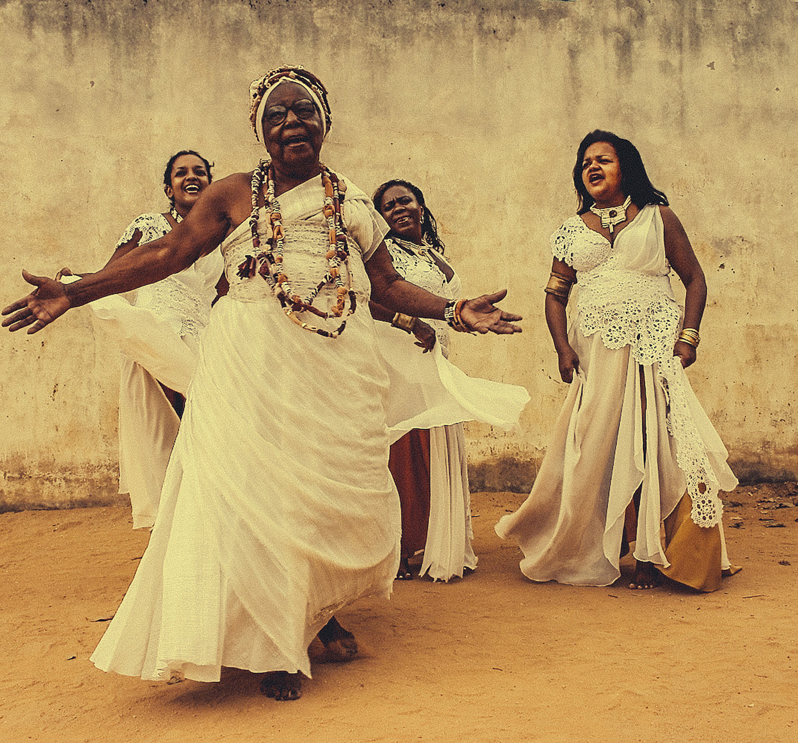 Quatro mulheres vestidas com trajes tradicionais brancos e colares de mi&ccedil;angas, cantando e dan&ccedil;ando ao ar livre.