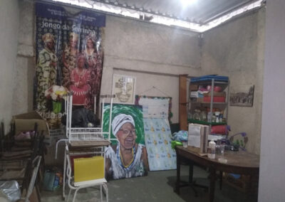 Um quarto desorganizado, com um grande pôster mostrando pessoas em trajes tradicionais, um quadro retratando uma senhora idosa e uma mesa com alguns objetos.