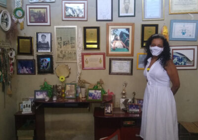 Uma mulher vestindo um vestido branco e uma máscara facial está em pé em uma sala com muitas fotos emolduradas, certificados e prêmios expostos na parede atrás dela.