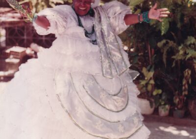 Uma pessoa sorridente vestindo um traje tradicional baiano branco, com saia larga, véu na cabeça e um grande adereço plano na cabeça, em pé com os braços estendidos.