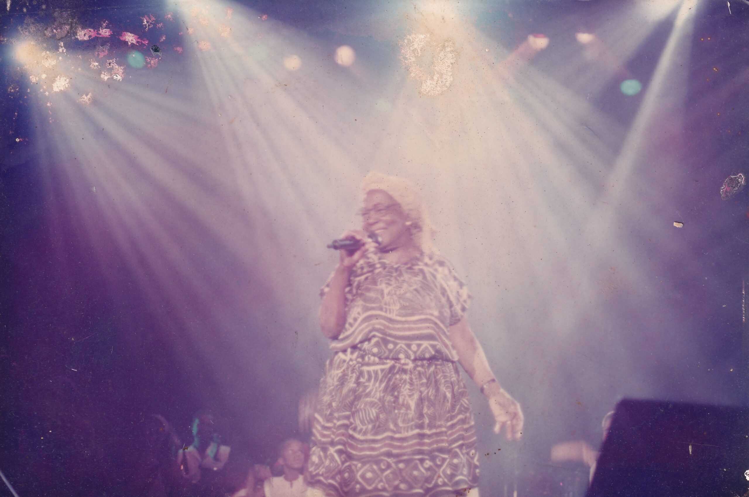 Uma mulher cantando ao microfone em um palco, sob holofotes intensos.