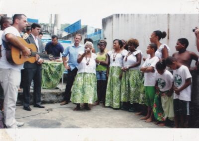 Um homem toca violão enquanto uma mulher mais velha canta no microfone, cercada por um grupo de adultos e crianças.