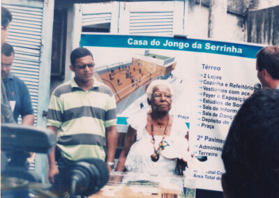 Uma senhora idosa e um homem estão em frente a um cartaz intitulado “Casa do Jongo da Serrinha”, que mostra uma planta baixa arquitetônica e uma lista de cômodos.