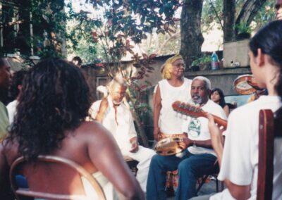 Um grupo de pessoas sentadas em círculo tocando música, incluindo um homem tocando violão e outro segurando um pandeiro.