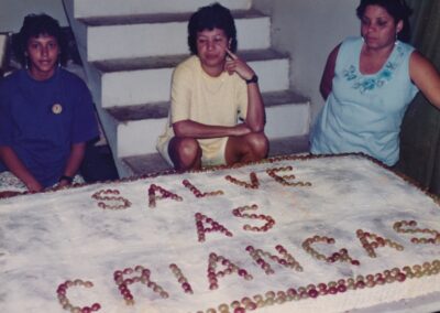 Três mulheres estão sentadas atrás de um grande bolo retangular decorado com uvas que formam a frase “SALVE AS CRIANÇAS”.