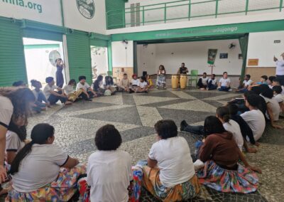 Um grande grupo de pessoas está sentado em círculo sobre um piso de pedra estampado para participar de uma oficina com dois músicos tocando tambores no centro.