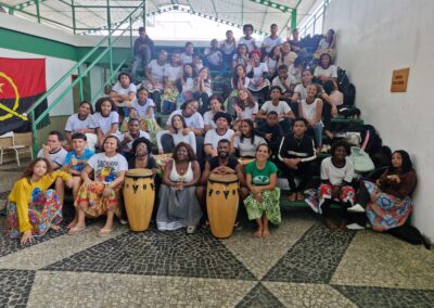 Um grande grupo de pessoas sentadas em fileiras nas arquibancadas de concreto, com duas pessoas na frente segurando grandes tambores djembe.